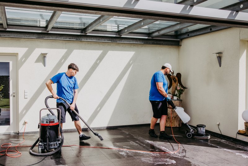 Zwei Mitarbeiter von cleanteam reinigen den Boden einer überdachten Terrasse mit Nasssauger und Einscheibenmaschine